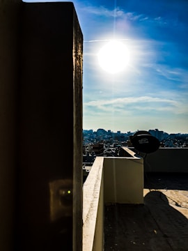 The image captures a bright sun in a clear blue sky above an urban landscape. In the foreground, there is a rooftop area with a dish antenna. Shadows are cast on the wall and floor of the rooftop from the sun's position.