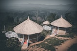Two circular, thatched-roof huts with raised wooden platforms and small stairs are nestled in a lush, mountainous landscape. The huts are surrounded by greenery, with distant hills visible in the background under a subdued sky.