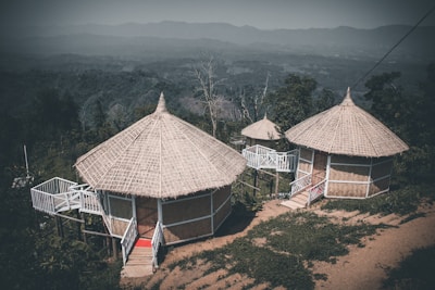 Two circular, thatched-roof huts with raised wooden platforms and small stairs are nestled in a lush, mountainous landscape. The huts are surrounded by greenery, with distant hills visible in the background under a subdued sky.