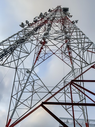 A tall metal communication tower reaching into the sky, featuring a complex lattice of red and white structural elements. Various antennas and satellite dishes are attached at different levels, indicating its use for broadcasting or telecommunications.