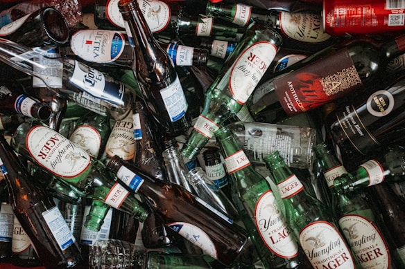 A large collection of empty beer and wine bottles scattered in various orientations. The labels on the bottles are prominently visible, with brands such as Yuengling, Michelob Ultra, and Corona Extra. The bottles vary in size and are primarily green, brown, or clear glass.