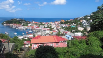 A picturesque coastal town with vibrant, colorful buildings surrounded by lush greenery. The bright blue sea stretches into the distance, dotted with small boats, while the sky is clear with a few scattered clouds.