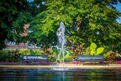 A serene park setting with a water fountain in the center, surrounded by lush green trees and plants. Two benches face the fountain, one with the words 'Spartans Will Not Be Bystanders' inscribed on it. Bright pink flowers line the garden area, and a brick building is partially visible in the background.