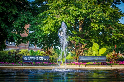 A serene park setting with a water fountain in the center, surrounded by lush green trees and plants. Two benches face the fountain, one with the words 'Spartans Will Not Be Bystanders' inscribed on it. Bright pink flowers line the garden area, and a brick building is partially visible in the background.