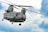 A large military helicopter is flying against a partly cloudy sky. The helicopter is grey-green in color and has tandem rotors. There are multiple windows along its side and various equipment attached to its exterior.