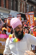 A person wearing a pink turban and a white shirt is in the foreground, adorned with a colorful rainbow lei. Around them are people holding signs that read 'NDP' and waving rainbow flags, indicating participation in a parade. The background shows a lively crowd and buildings with various storefronts.