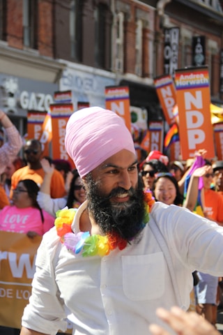 A person wearing a pink turban and a white shirt is in the foreground, adorned with a colorful rainbow lei. Around them are people holding signs that read 'NDP' and waving rainbow flags, indicating participation in a parade. The background shows a lively crowd and buildings with various storefronts.