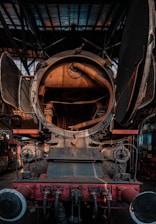 The image captures the inside of a vintage steam locomotive. The front view of the engine's interior showcases mechanical components including a large circular opening, possibly the firebox or boiler, and various pipes and fittings. The metallic surface has a rustic, aged look with visible signs of wear and rust. Industrial surroundings are visible, indicating a workshop or train shed setting.