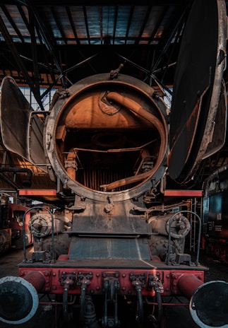 The image captures the inside of a vintage steam locomotive. The front view of the engine's interior showcases mechanical components including a large circular opening, possibly the firebox or boiler, and various pipes and fittings. The metallic surface has a rustic, aged look with visible signs of wear and rust. Industrial surroundings are visible, indicating a workshop or train shed setting.