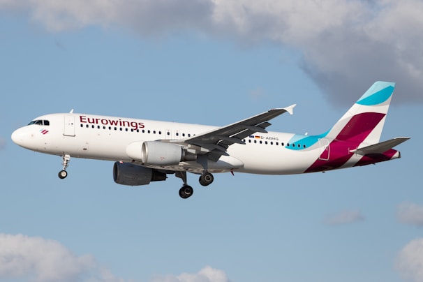 An airplane with Eurowings branding is flying in a clear sky. The aircraft is white with blue and magenta accents and is visible with landing gear down.