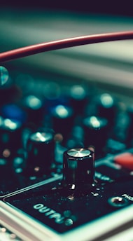Various knobs and controls on an electronic audio device are in focus, with a red wire prominently curving across the frame. The device displays settings and has a chrome-like finish, reflecting light.