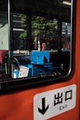 A view inside a bus or train with an orange interior. A blue ticket machine is visible next to a window showing a reflection of trees and buildings outside. An exit sign with Japanese characters is prominent in the foreground.