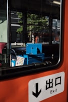 A view inside a bus or train with an orange interior. A blue ticket machine is visible next to a window showing a reflection of trees and buildings outside. An exit sign with Japanese characters is prominent in the foreground.