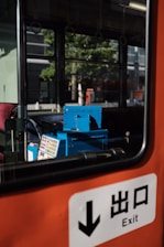 A view inside a bus or train with an orange interior. A blue ticket machine is visible next to a window showing a reflection of trees and buildings outside. An exit sign with Japanese characters is prominent in the foreground.