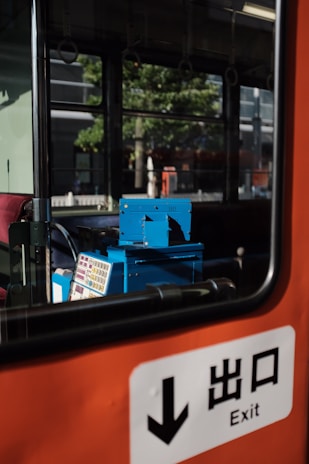 A view inside a bus or train with an orange interior. A blue ticket machine is visible next to a window showing a reflection of trees and buildings outside. An exit sign with Japanese characters is prominent in the foreground.