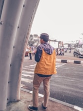 A person wearing a yellow jacket and beige pants stands at the edge of a crosswalk, looking towards a street filled with traffic. Several vehicles, including motorcycles, are visible on the road. The background features urban buildings and a few trees. A police officer directs traffic, adding a sense of bustle to the scene.