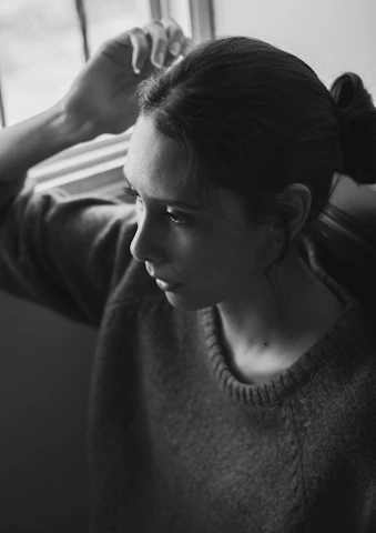 A cinematic-style photo of a mature woman looking thoughtfully out a window, soft beige and rose tones surrounding her.