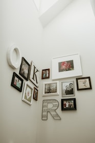 A collection of framed photographs and artworks is arranged on a light-colored wall. The display includes various sizes and styles of frames, featuring family photos, portraits, and decorative letters 'K' and 'R'. The images include a flower, a forest scene, and people in different poses.