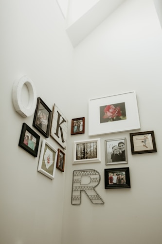 A collection of framed photographs and artworks is arranged on a light-colored wall. The display includes various sizes and styles of frames, featuring family photos, portraits, and decorative letters 'K' and 'R'. The images include a flower, a forest scene, and people in different poses.