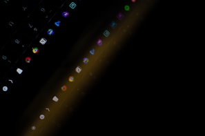A dimly lit laptop keyboard is partially visible with illuminated app icons displayed on the screen. The image captures a blurred reflection of these icons across the keyboard surface, with vibrant colors contrasting against a predominantly dark background.