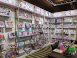 A retail display featuring a wide array of colorful children's craft and activity kits arranged on shelves. The kits are packed in vibrant boxes showcasing activities like jewelry making and crafting. A table in the foreground is adorned with neon-colored water bottles, crafting supplies, and a bright pink bag.