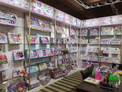 A retail display featuring a wide array of colorful children's craft and activity kits arranged on shelves. The kits are packed in vibrant boxes showcasing activities like jewelry making and crafting. A table in the foreground is adorned with neon-colored water bottles, crafting supplies, and a bright pink bag.