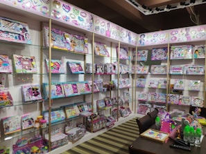 A retail display featuring a wide array of colorful children's craft and activity kits arranged on shelves. The kits are packed in vibrant boxes showcasing activities like jewelry making and crafting. A table in the foreground is adorned with neon-colored water bottles, crafting supplies, and a bright pink bag.