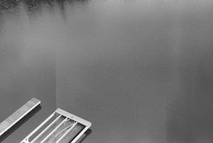 A black and white photograph showing a serene body of water with a wooden dock or pier extending into it. The calm water reflects textures and shadows, likely of nearby surroundings. The composition is minimalistic with a focus on the contrast between the smooth water surface and the linear structure of the dock.