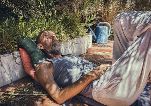 A man is lying on the ground with his eyes closed, resting his head on a green pillow and red cushion. He is surrounded by greenery and plants, with a blue watering can nearby on the brick surface. Shadows of leaves create patterns on his body, and he appears relaxed.