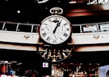 A large, ornate clock with multiple dials hangs in a shopping center. Roman numerals adorn the main face, while smaller dials track different time zones. The clock's design is intricate, featuring gears and mechanical elements. Holiday decorations are visible in the background.