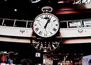 A large, ornate clock with multiple dials hangs in a shopping center. Roman numerals adorn the main face, while smaller dials track different time zones. The clock's design is intricate, featuring gears and mechanical elements. Holiday decorations are visible in the background.