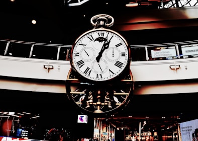 A large, ornate clock with multiple dials hangs in a shopping center. Roman numerals adorn the main face, while smaller dials track different time zones. The clock's design is intricate, featuring gears and mechanical elements. Holiday decorations are visible in the background.