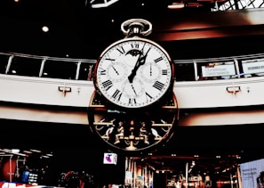 A large, ornate clock with multiple dials hangs in a shopping center. Roman numerals adorn the main face, while smaller dials track different time zones. The clock's design is intricate, featuring gears and mechanical elements. Holiday decorations are visible in the background.