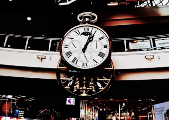 A large, ornate clock with multiple dials hangs in a shopping center. Roman numerals adorn the main face, while smaller dials track different time zones. The clock's design is intricate, featuring gears and mechanical elements. Holiday decorations are visible in the background.