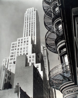 A black-and-white photograph captures an urban scene with tall skyscrapers and intricate, curved fire escapes along the side of a building. Shadows and light play off the geometric patterns, creating a striking contrast.