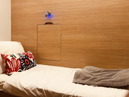 A minimalist wooden wall with a small blue-lit fixture above a recessed panel. In front of the wall is a simple white bed with a colorful floral pillow and a folded grey blanket.