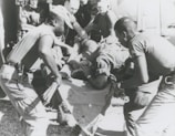 A group of individuals are engaged in moving or assisting a person on a stretcher. The scene appears to involve military personnel, possibly during a rescue or emergency situation. The environment suggests they might be near a vehicle or aircraft.