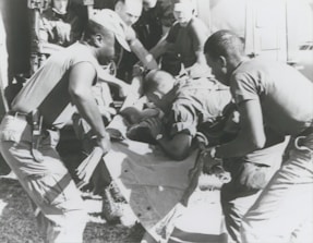 A group of individuals are engaged in moving or assisting a person on a stretcher. The scene appears to involve military personnel, possibly during a rescue or emergency situation. The environment suggests they might be near a vehicle or aircraft.