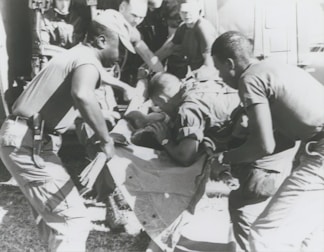 A group of individuals are engaged in moving or assisting a person on a stretcher. The scene appears to involve military personnel, possibly during a rescue or emergency situation. The environment suggests they might be near a vehicle or aircraft.
