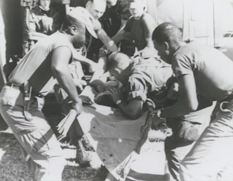 A group of individuals are engaged in moving or assisting a person on a stretcher. The scene appears to involve military personnel, possibly during a rescue or emergency situation. The environment suggests they might be near a vehicle or aircraft.
