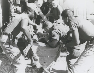 A group of individuals are engaged in moving or assisting a person on a stretcher. The scene appears to involve military personnel, possibly during a rescue or emergency situation. The environment suggests they might be near a vehicle or aircraft.