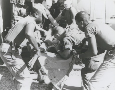 A group of individuals are engaged in moving or assisting a person on a stretcher. The scene appears to involve military personnel, possibly during a rescue or emergency situation. The environment suggests they might be near a vehicle or aircraft.
