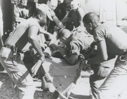 A group of individuals are engaged in moving or assisting a person on a stretcher. The scene appears to involve military personnel, possibly during a rescue or emergency situation. The environment suggests they might be near a vehicle or aircraft.