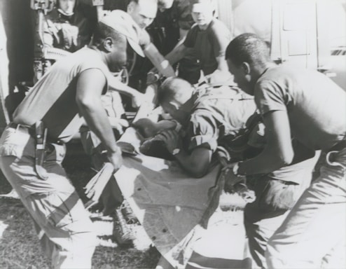 A group of individuals are engaged in moving or assisting a person on a stretcher. The scene appears to involve military personnel, possibly during a rescue or emergency situation. The environment suggests they might be near a vehicle or aircraft.
