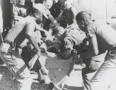 A group of individuals are engaged in moving or assisting a person on a stretcher. The scene appears to involve military personnel, possibly during a rescue or emergency situation. The environment suggests they might be near a vehicle or aircraft.