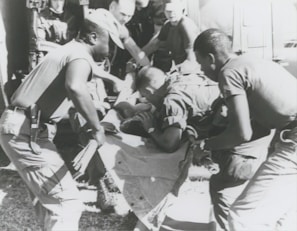 A group of individuals are engaged in moving or assisting a person on a stretcher. The scene appears to involve military personnel, possibly during a rescue or emergency situation. The environment suggests they might be near a vehicle or aircraft.