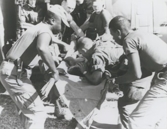 A group of individuals are engaged in moving or assisting a person on a stretcher. The scene appears to involve military personnel, possibly during a rescue or emergency situation. The environment suggests they might be near a vehicle or aircraft.