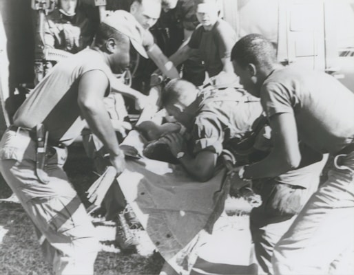 A group of individuals are engaged in moving or assisting a person on a stretcher. The scene appears to involve military personnel, possibly during a rescue or emergency situation. The environment suggests they might be near a vehicle or aircraft.
