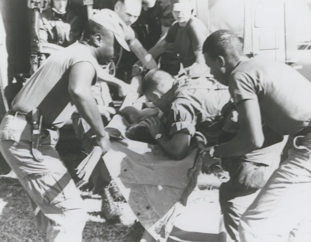 A group of individuals are engaged in moving or assisting a person on a stretcher. The scene appears to involve military personnel, possibly during a rescue or emergency situation. The environment suggests they might be near a vehicle or aircraft.