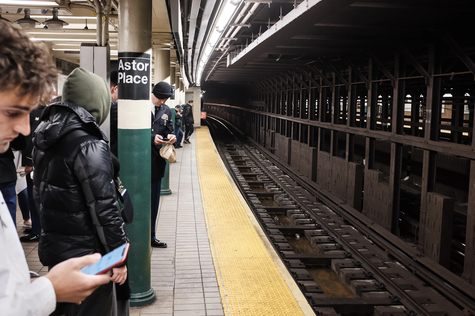 Riders waiting along the yellow edge strip of an underground platform, the tracks curving away into the tunnel
